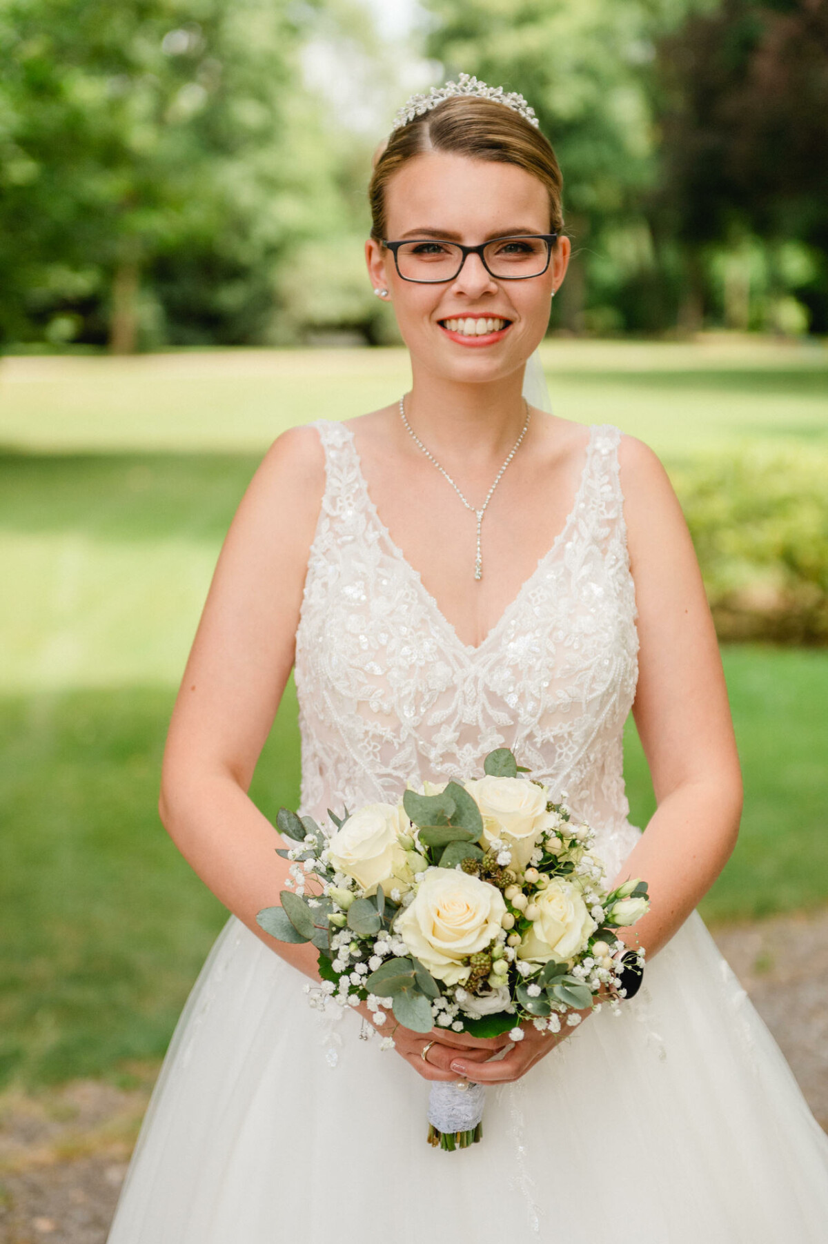 Eine Braut im weißen Spitzenkleid steht im Freien, hält einen Strauß weißer Rosen und Grünpflanzen in der Hand und lächelt in die Kamera.