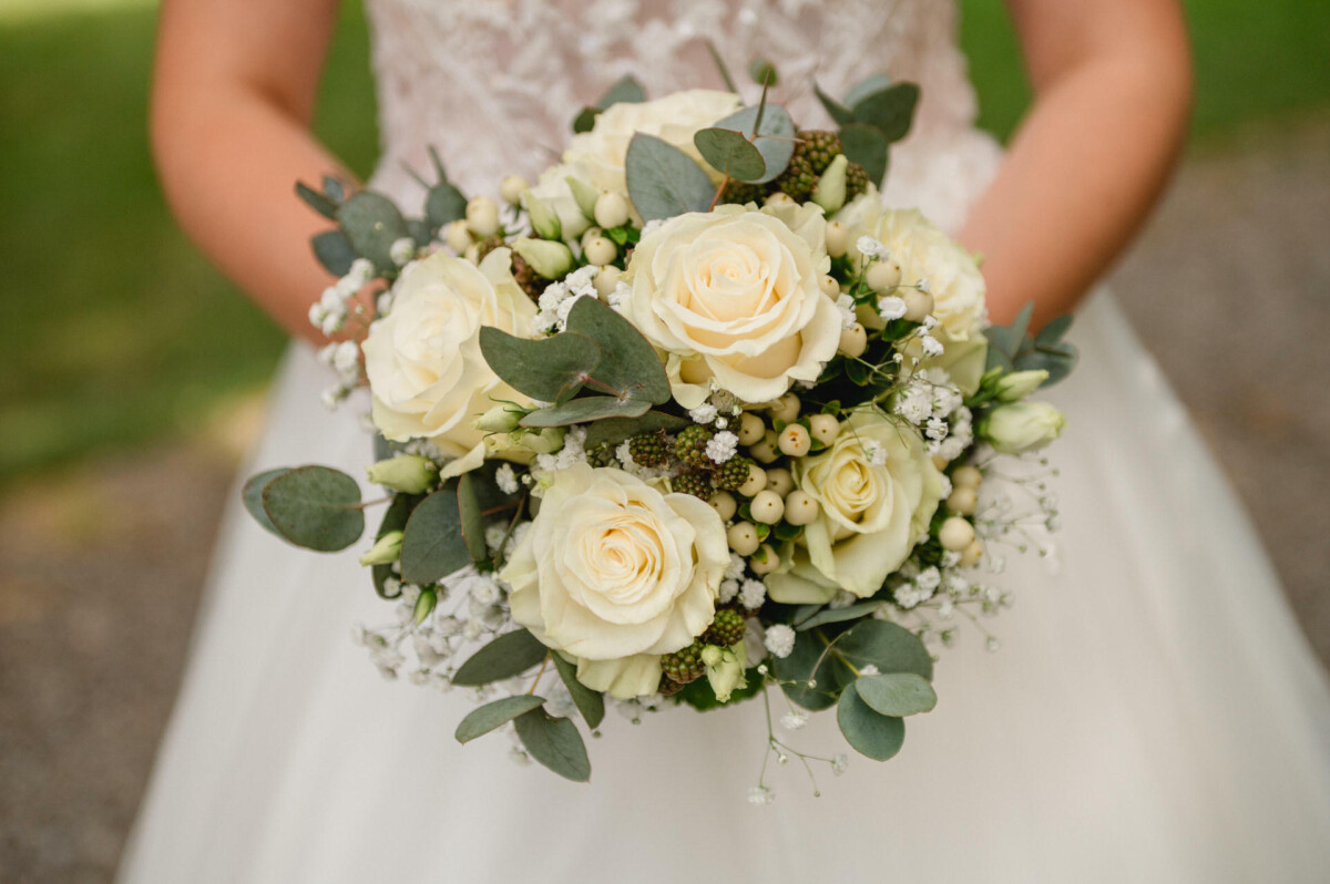 Eine Person hält einen Strauß weißer Rosen und Grünpflanzen und trägt ein weißes Spitzenkleid.