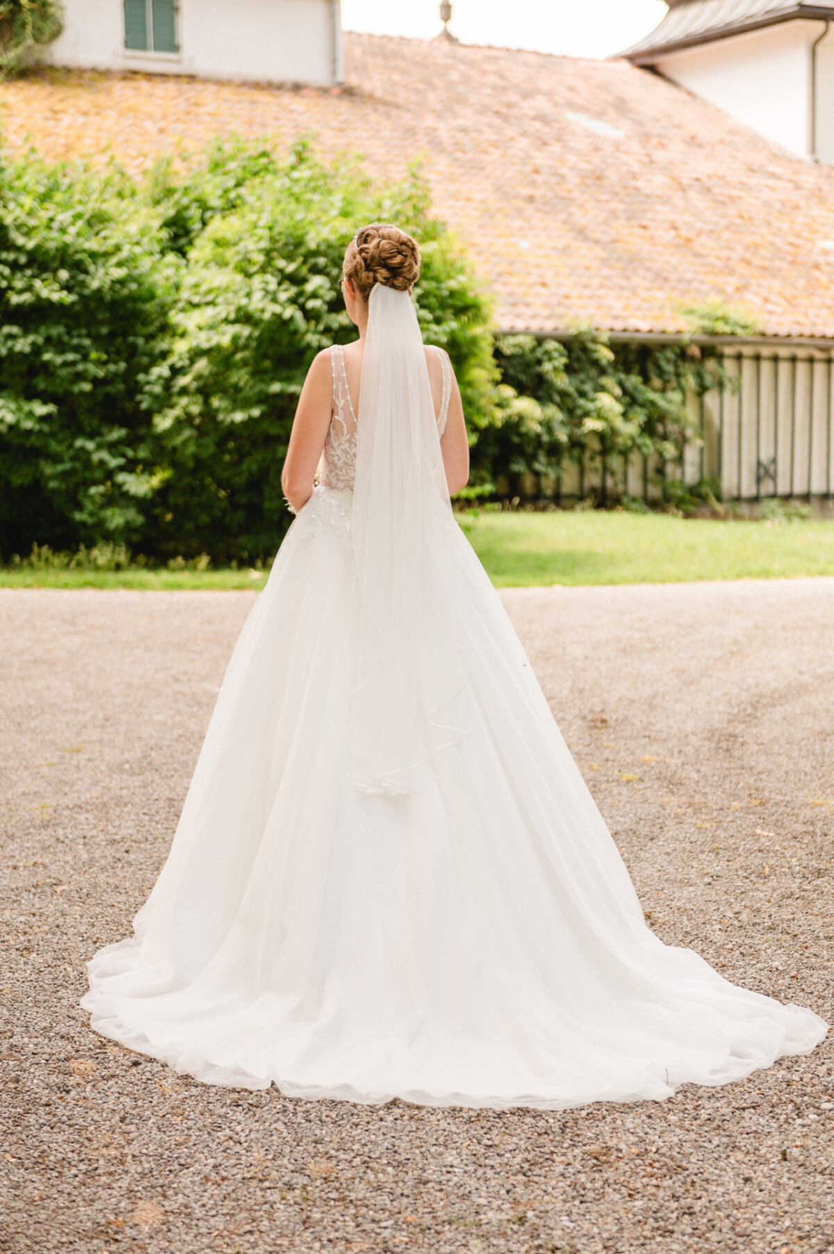 Eine Braut in einem weißen Kleid mit langer Schleppe und Schleier steht mit dem Rücken zur Kamera auf einem Kiesweg, im Hintergrund sind eine belaubte Kulisse und ein Hausdach zu sehen.