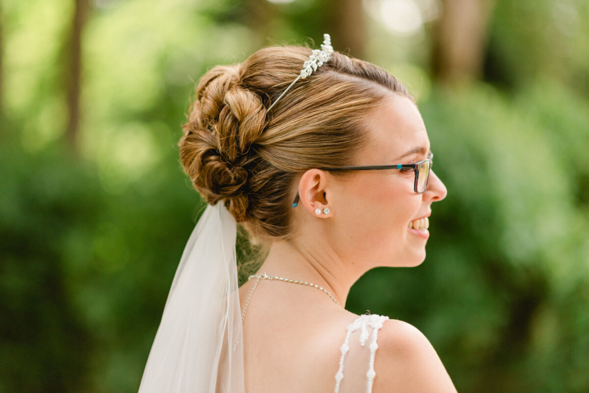 Eine Frau mit Brautfrisur und Schleier, die eine Brille und Ohrringe trägt, lächelt, während sie im Freien steht.