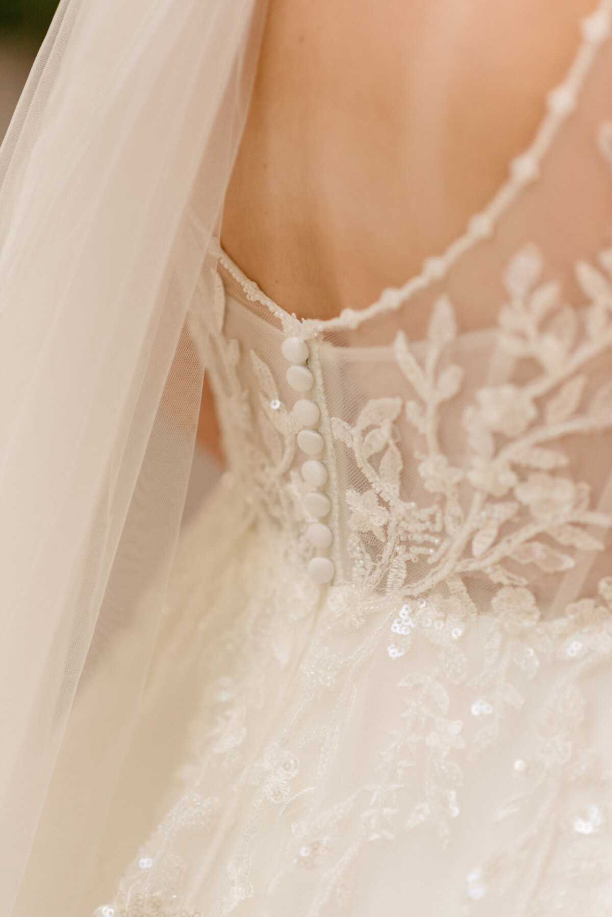 Eine Nahaufnahme eines Brautkleides mit aufwendiger Spitzenstickerei und kleinen Knöpfen auf transparentem Stoff. In der oberen linken Ecke ist ein Schleier sichtbar.