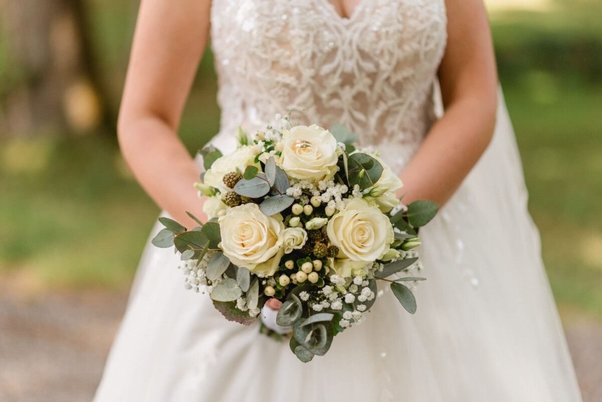 Eine Person in einem weißen Spitzenbrautkleid hält einen Strauß weißer Rosen und Grünpflanzen.