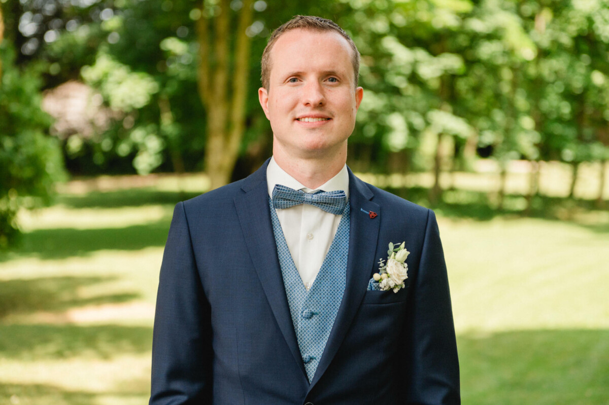 Ein Mann in einem marineblauen Anzug und einer Fliege steht draußen in einer grünen Parkanlage.