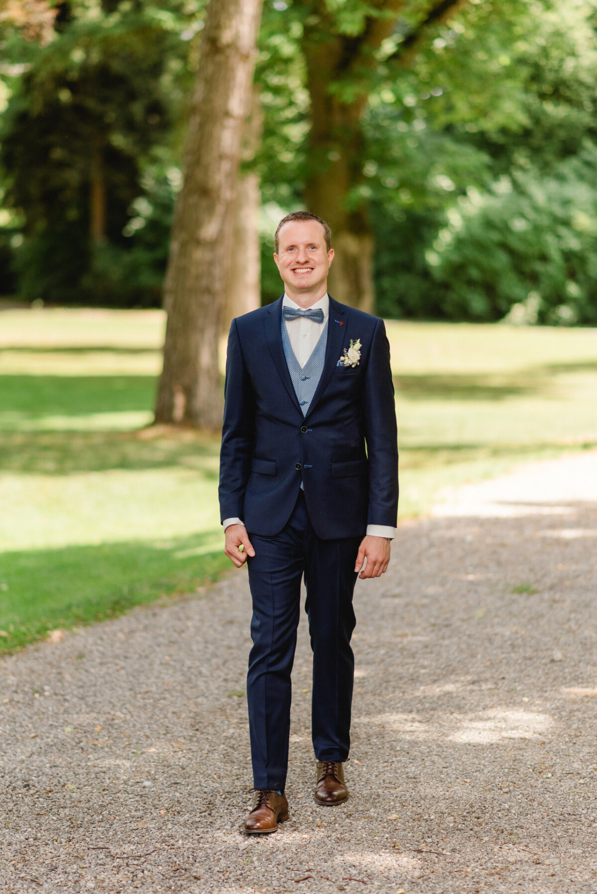 Ein Mann in einem marineblauen Anzug mit Fliege steht auf einem Kiesweg, umgeben von Bäumen und grünem Gras.