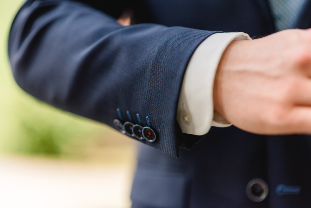 Nahaufnahme einer Person, die einen dunklen Anzug mit sichtbaren Manschettenknöpfen und einem weißen Hemd darunter trägt und vor einem unscharfen Hintergrund ihren Ärmel zurechtrückt.