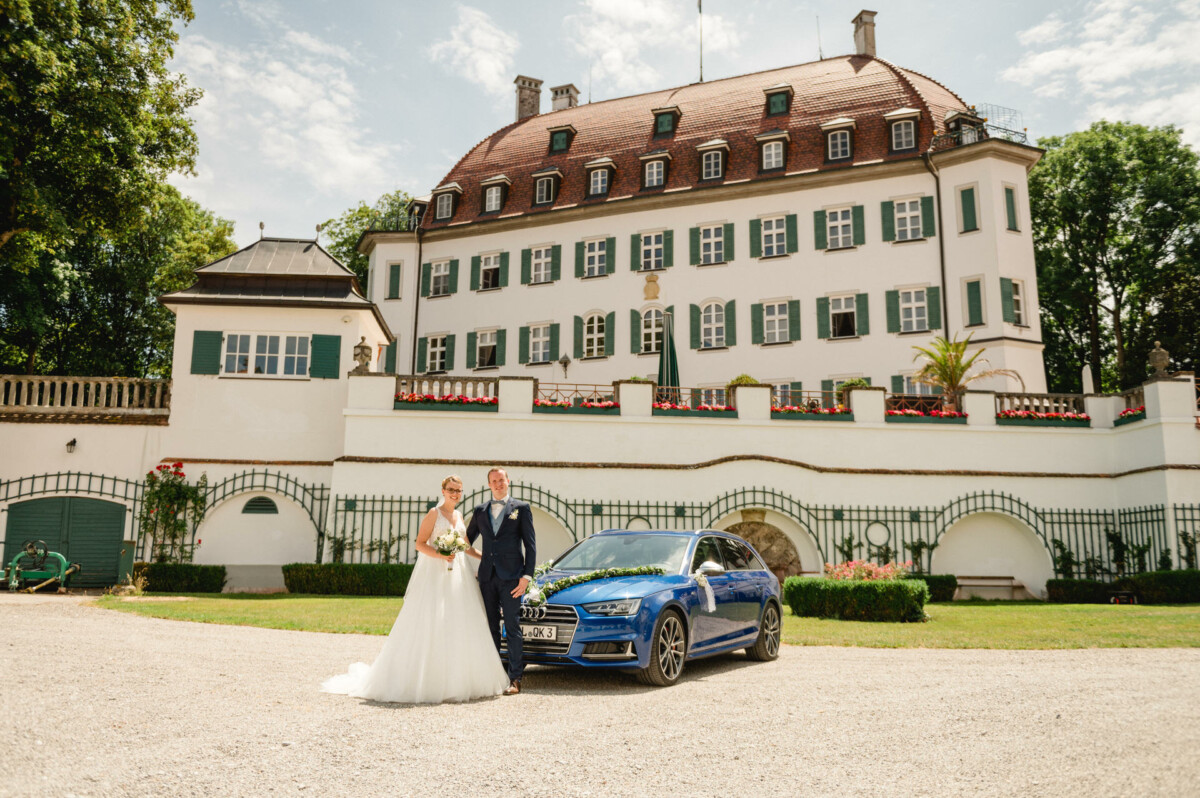 Braut und Bräutigam stehen neben einem blauen Auto vor einem großen, reich verzierten Gebäude.