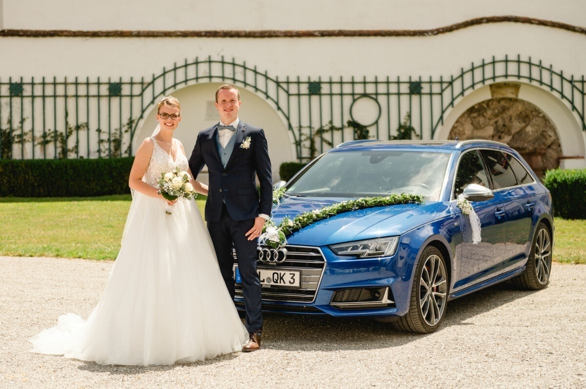 Braut und Bräutigam stehen neben einem mit Blumen geschmückten blauen Auto.