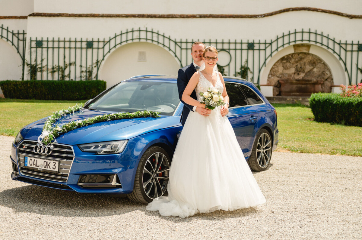 Braut und Bräutigam posieren vor einem mit Blumen geschmückten blauen Audi.