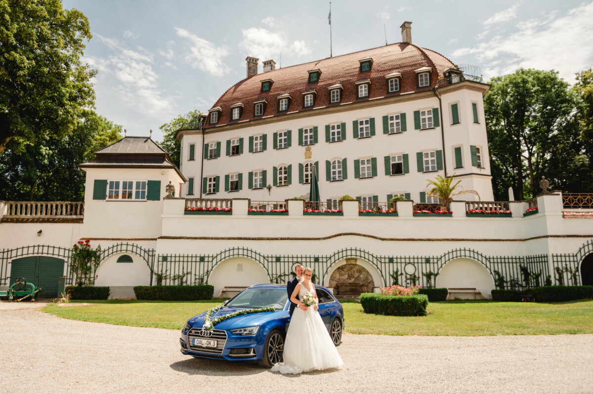 An einem sonnigen Tag posieren Braut und Bräutigam neben einem blauen Auto vor einem großen weißen Herrenhaus mit rotem Dach.