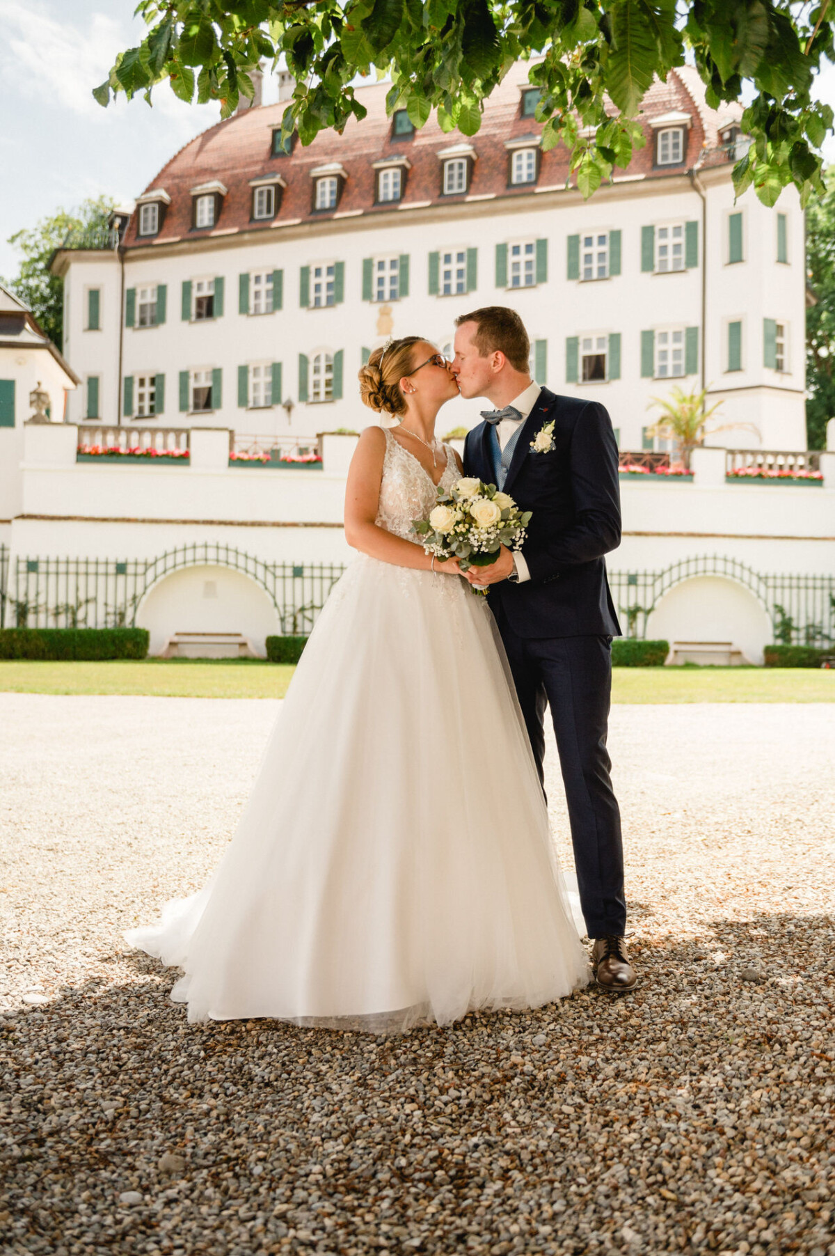 Braut und Bräutigam küssen sich vor einem großen, eleganten Gebäude, umgeben von Grün. Die Braut trägt ein weißes Kleid und der Bräutigam einen dunklen Anzug.