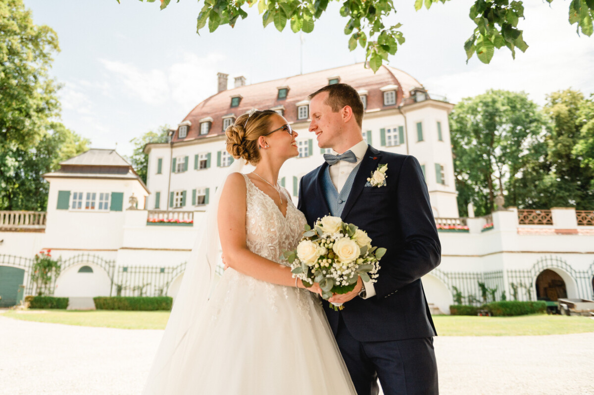 Braut und Bräutigam stehen mit einem Blumenstrauß in der Hand im Freien vor einem großen, eleganten Gebäude, umgeben von Grün.