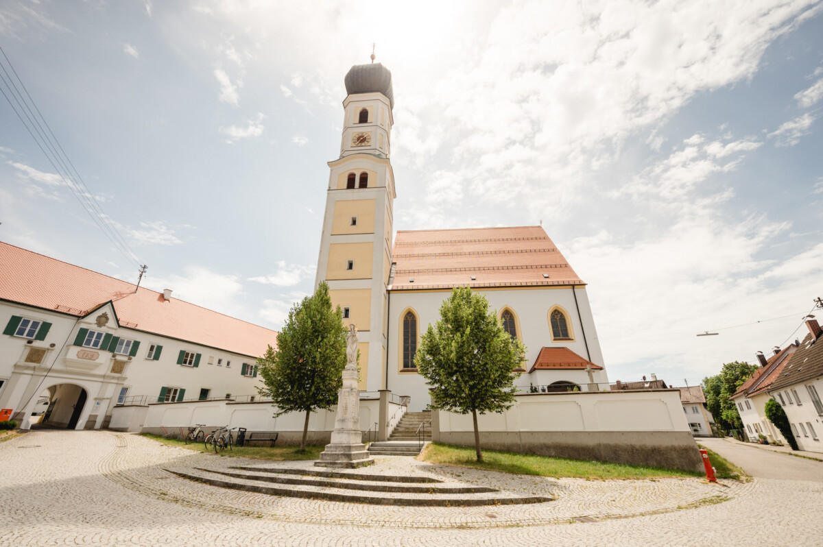 Eine sonnenbeschienene Kirche mit einem hohen, gewölbten Kirchturm steht neben einem Kopfsteinpflasterweg, gesäumt von Bäumen und umgeben von angrenzenden Gebäuden unter einem teilweise bewölkten Himmel.