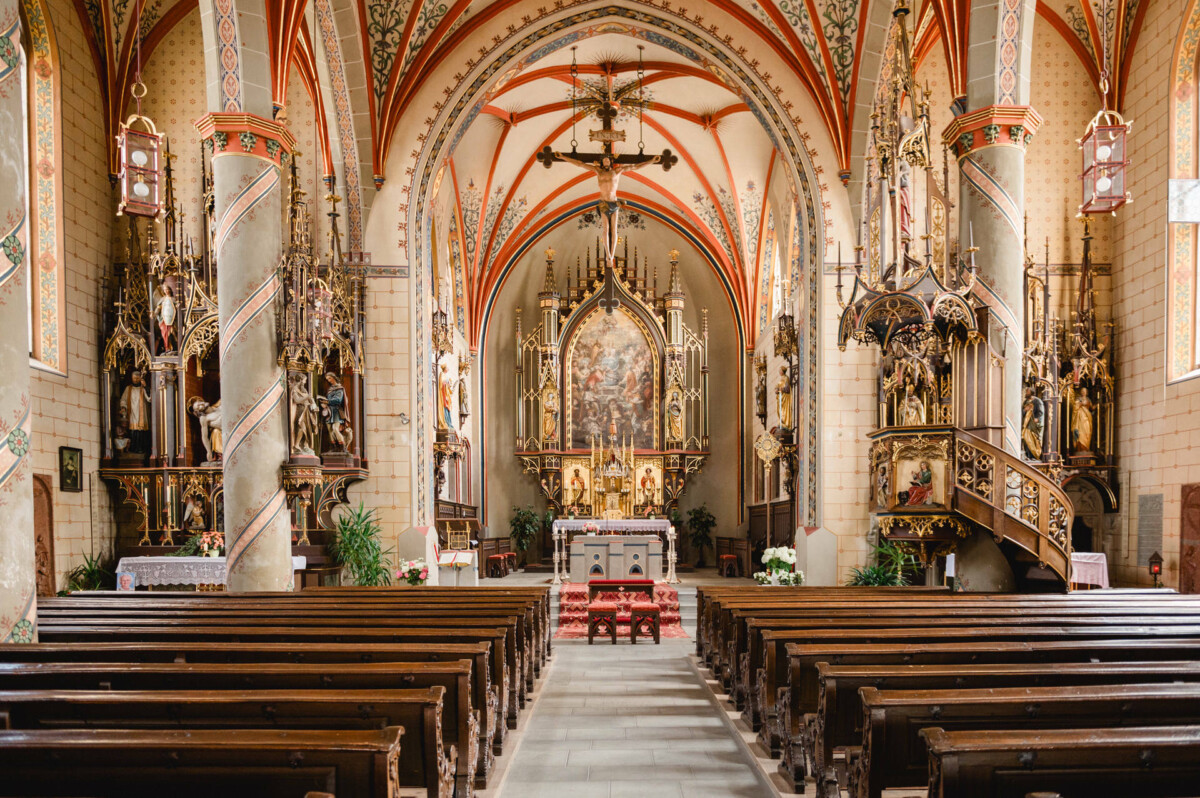 Innenansicht einer Kirche mit verzierten Säulen, dekorativen Bögen und einem detailreichen Altar. Holzbänke säumen den Gang, der zum Altar führt.