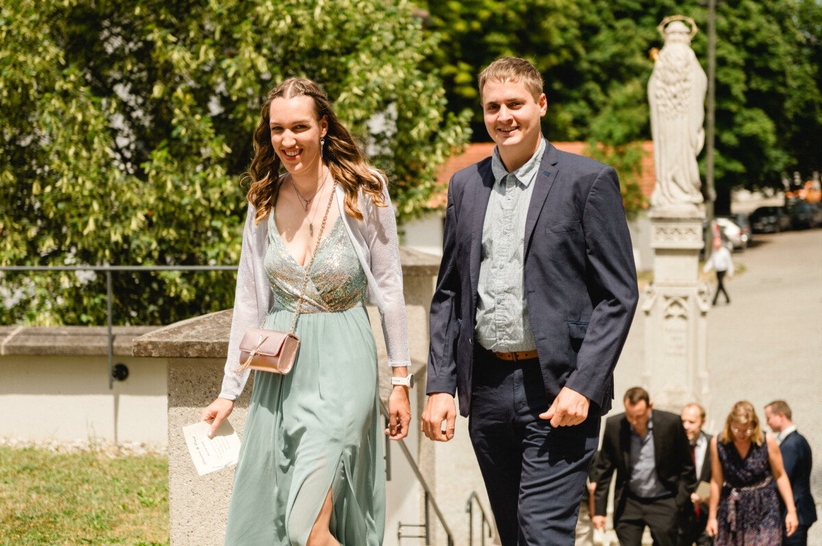 Eine Frau in einem grünen Kleid und ein Mann im Anzug gehen eine Außentreppe hinauf. Im Hintergrund ist eine Gruppe von Menschen in der Nähe einer Statue zu sehen.
