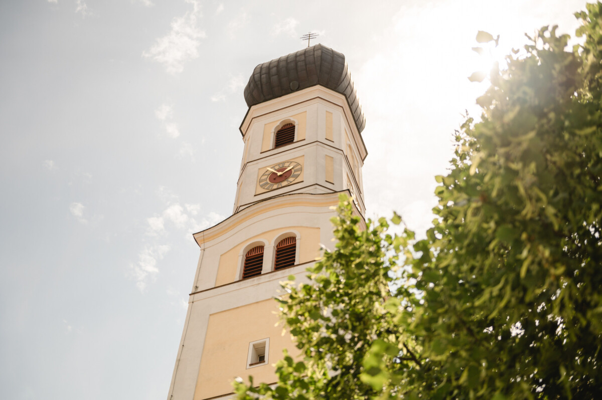 Hoher Uhrenturm mit schwarzem Dach vor einem klaren Himmel, teilweise verdeckt von grünen Blättern.
