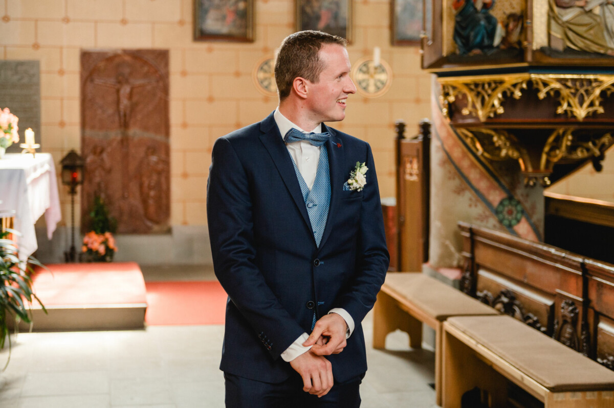 Ein Mann im blauen Anzug steht in einer Kirche und richtet mit neutralem Gesichtsausdruck seine Manschette.