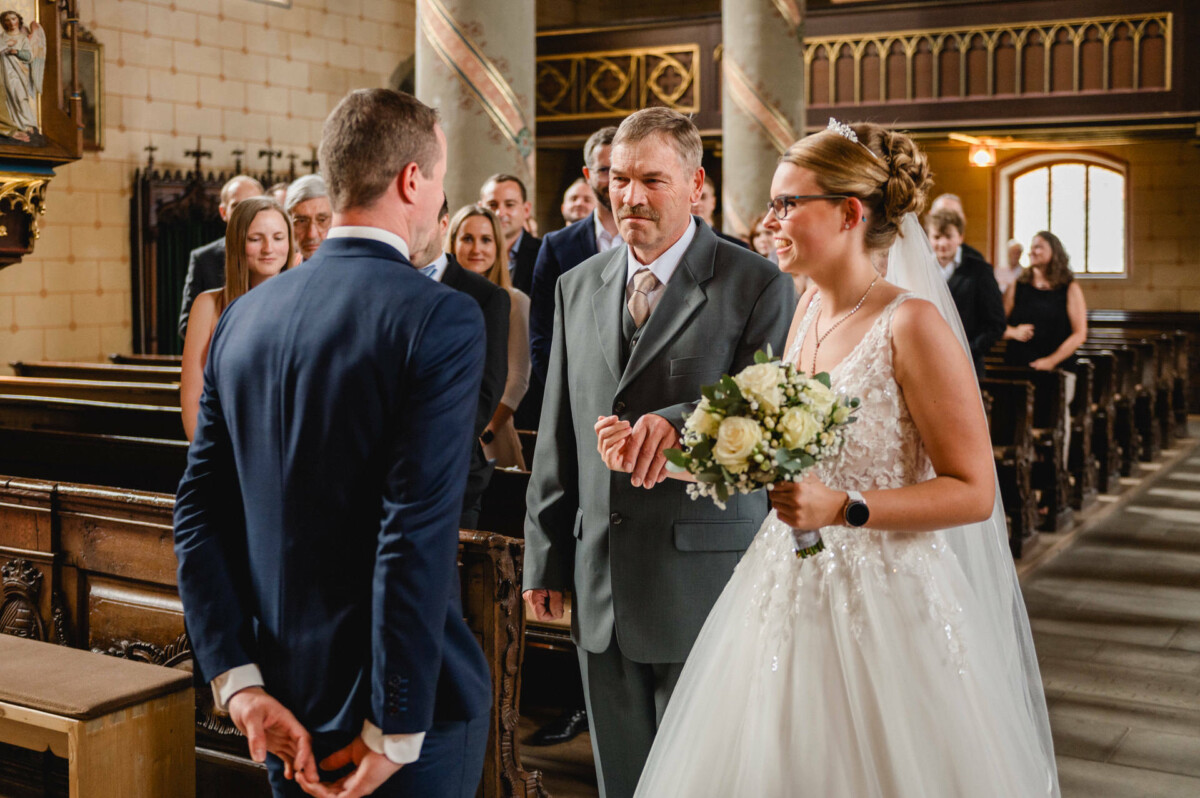 Eine Braut mit einem Blumenstrauß steht neben einem Mann im Anzug. Ihnen gegenüber steht ein weiterer Mann im marineblauen Anzug in einer Kirche. Im Hintergrund sitzen Gäste.