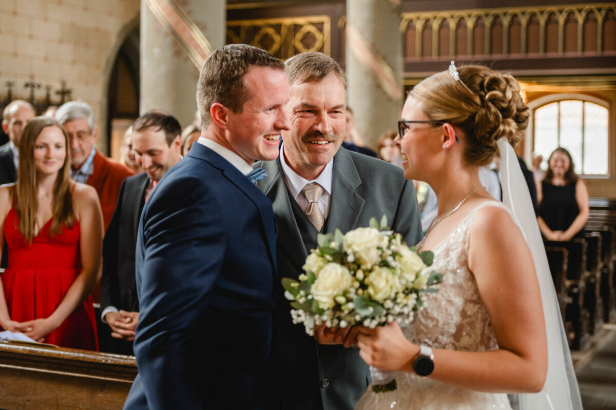 Eine Braut mit einem Blumenstrauß lächelt ihren Bräutigam und einen älteren Mann im Anzug in einer Kirche an, umgeben von sitzenden Gästen.