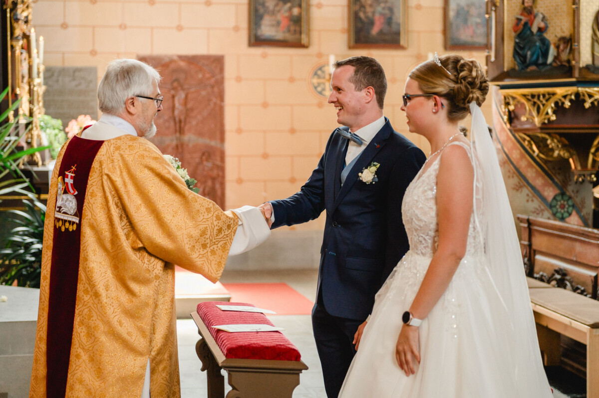 Ein Paar hält Händchen und lächelt einen Priester während einer Hochzeitszeremonie in einer Kirche an.