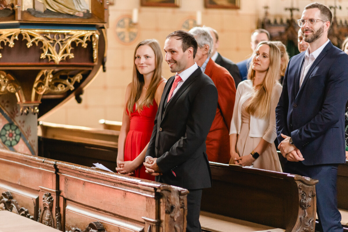 Eine Gruppe von Menschen steht und lächelt in einer Kirche. Man sieht Holzbänke und kunstvolle Verzierungen.