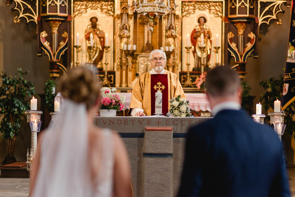 Ein Priester führt eine Hochzeitszeremonie an einem reich verzierten Altar durch, vor ihm stehen zwei Menschen.