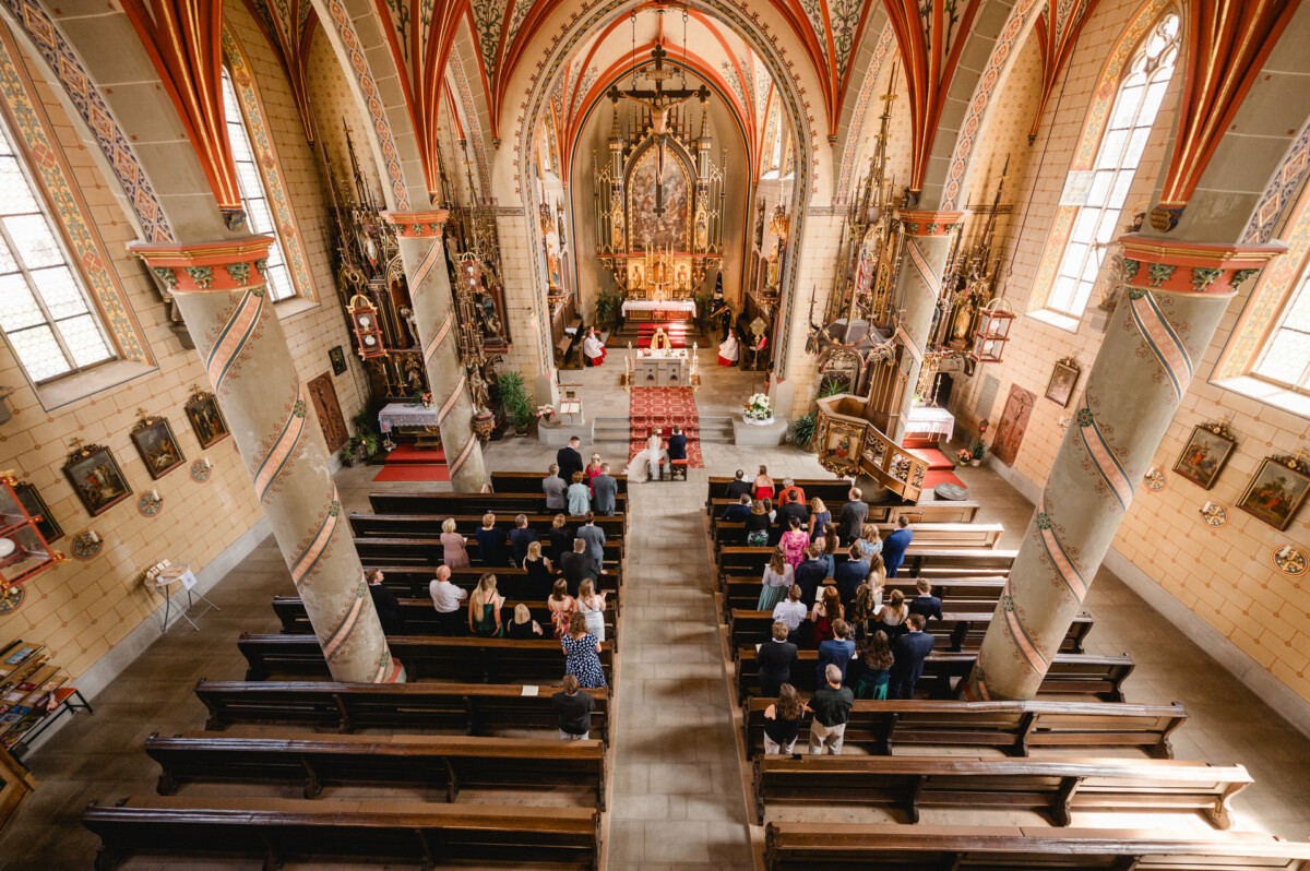 Innenansicht einer geschmückten Kirche mit Menschen, die während einer Zeremonie in Kirchenbänken sitzen und dem Altar zugewandt sind. Die Architektur zeichnet sich durch hohe Säulen und detaillierte Deckengestaltungen aus.