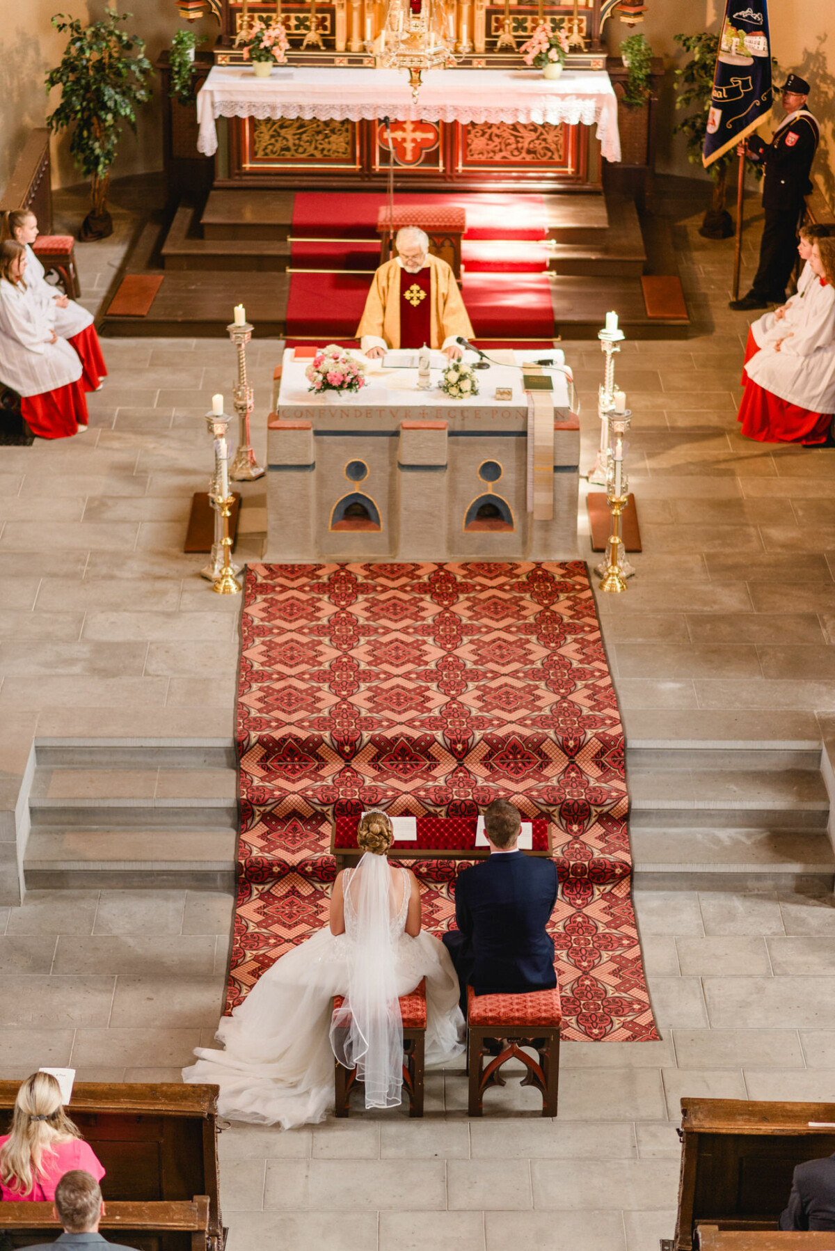 Ein Paar sitzt vor einem Altar in einer Kirche, gegenüber einem Geistlichen in Robe. Kerzen und Blumenschmuck sind vorhanden. Brautjungfern und Trauzeugen stehen in der Nähe.