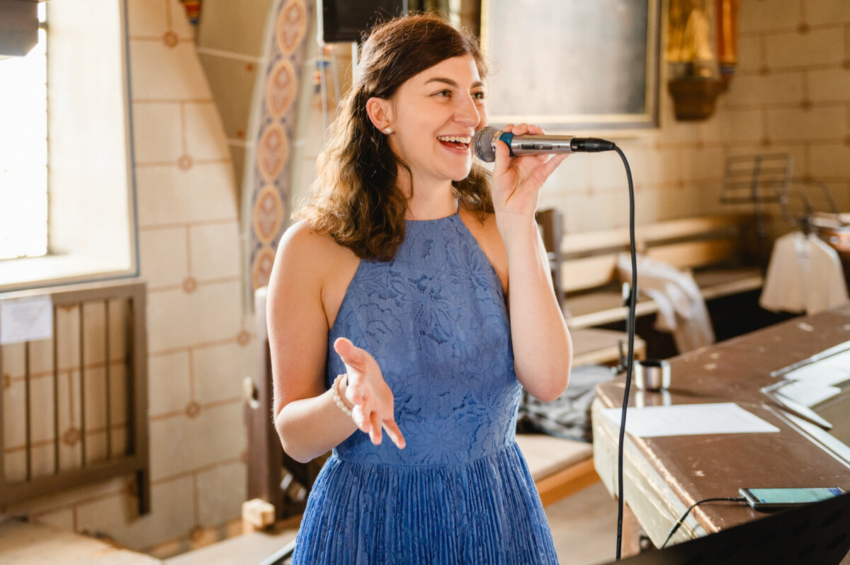 Eine Frau in einem blauen Kleid singt in einem Innenbereich neben einem Klavier in ein Mikrofon.