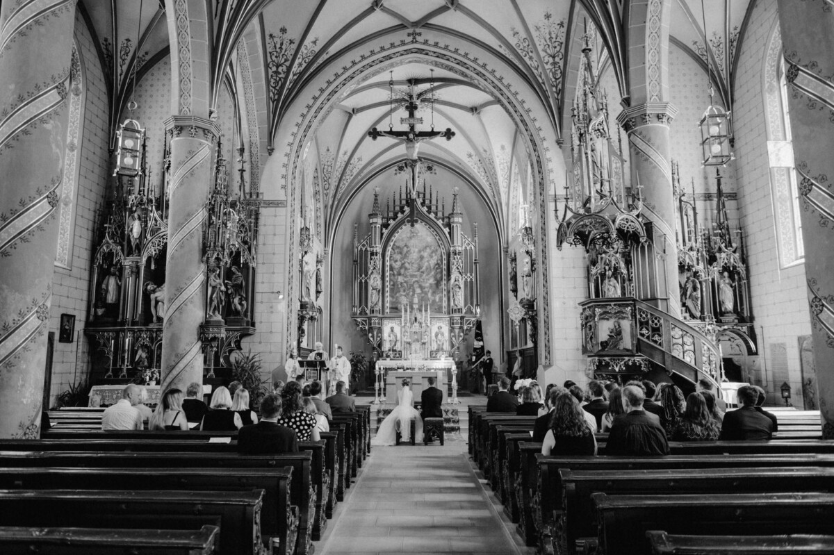 Eine Hochzeitszeremonie findet in einer Kirche im gotischen Stil statt. Die Gäste sitzen in den Kirchenbänken mit Blick auf den Altar.