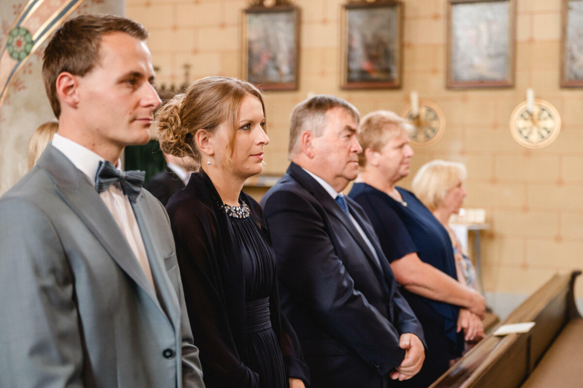 Eine Gruppe formell gekleideter Menschen steht in einer Kirche. An den Wänden hinter ihnen sind Gemälde zu sehen.