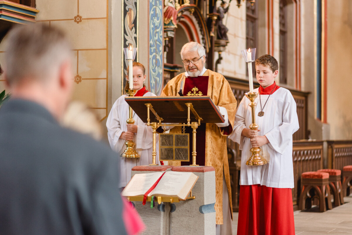In einer geschmückten Kirche steht ein Priester an einem Rednerpult und liest, flankiert von zwei Messdienern, die Kerzenleuchter halten.