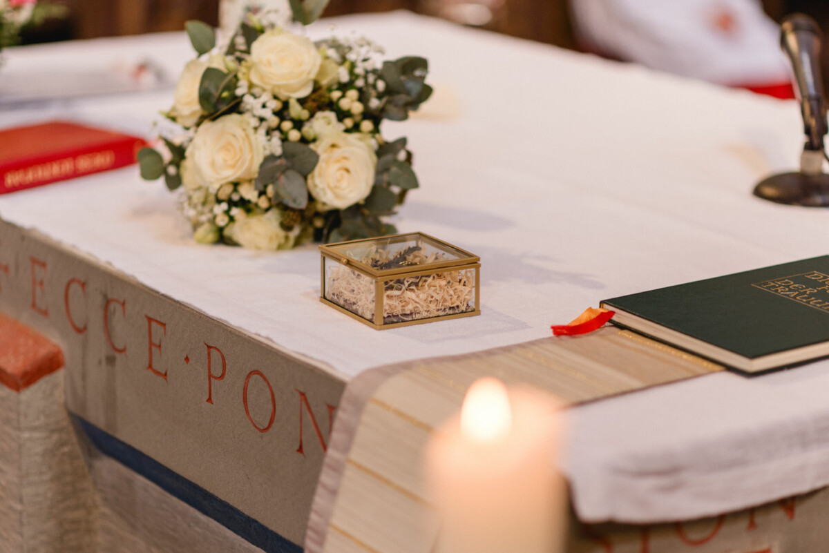 Ein Strauß weißer Rosen und eine kleine goldene Schachtel mit Asche stehen neben einem grünen Buch auf einem Kirchenaltar.