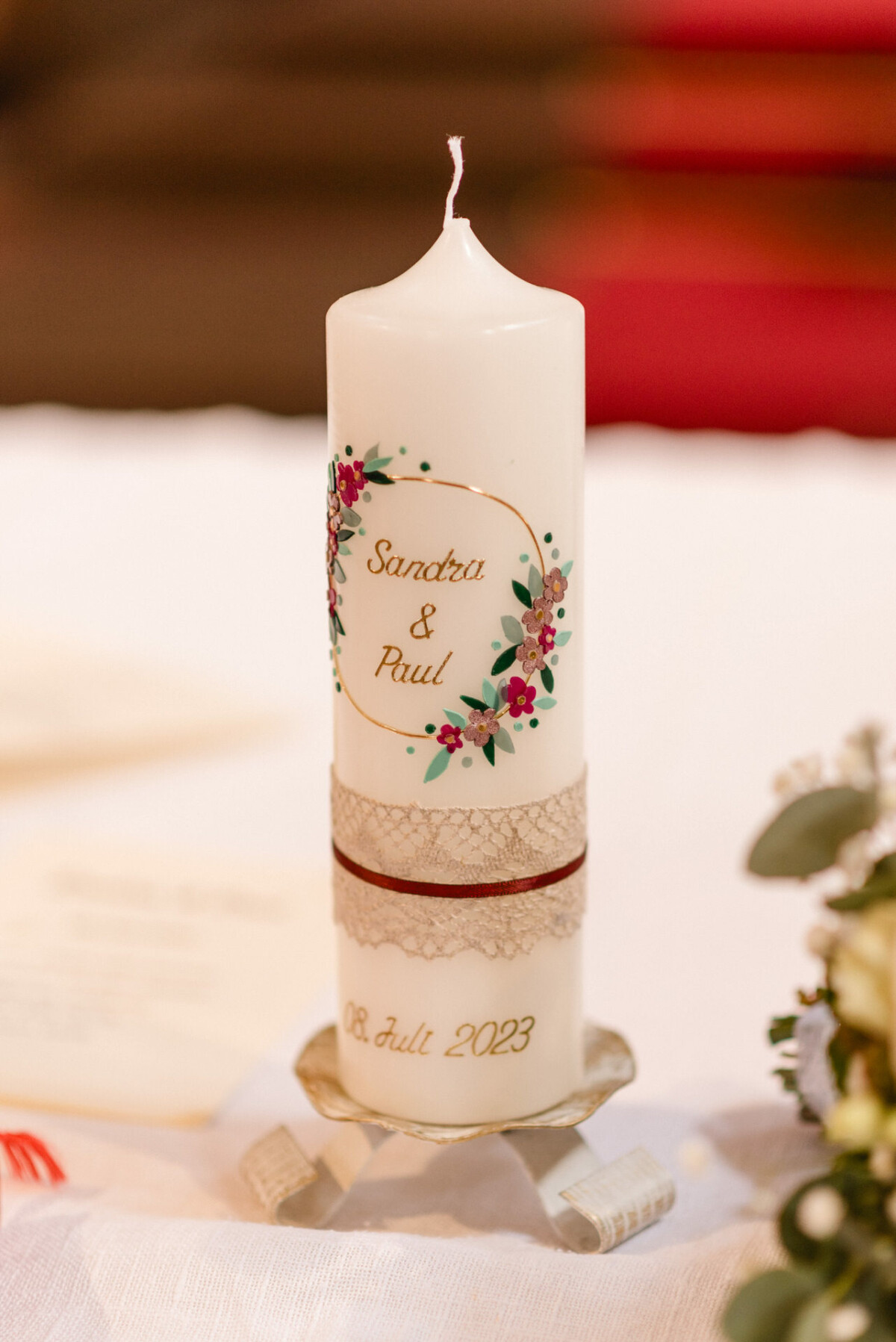 Eine dekorative Hochzeitskerze mit der Aufschrift „Sandra & Paul“ mit Blumenmuster und dem Datum „08. Juli 2023“, platziert auf einem Halter auf einem Tisch.