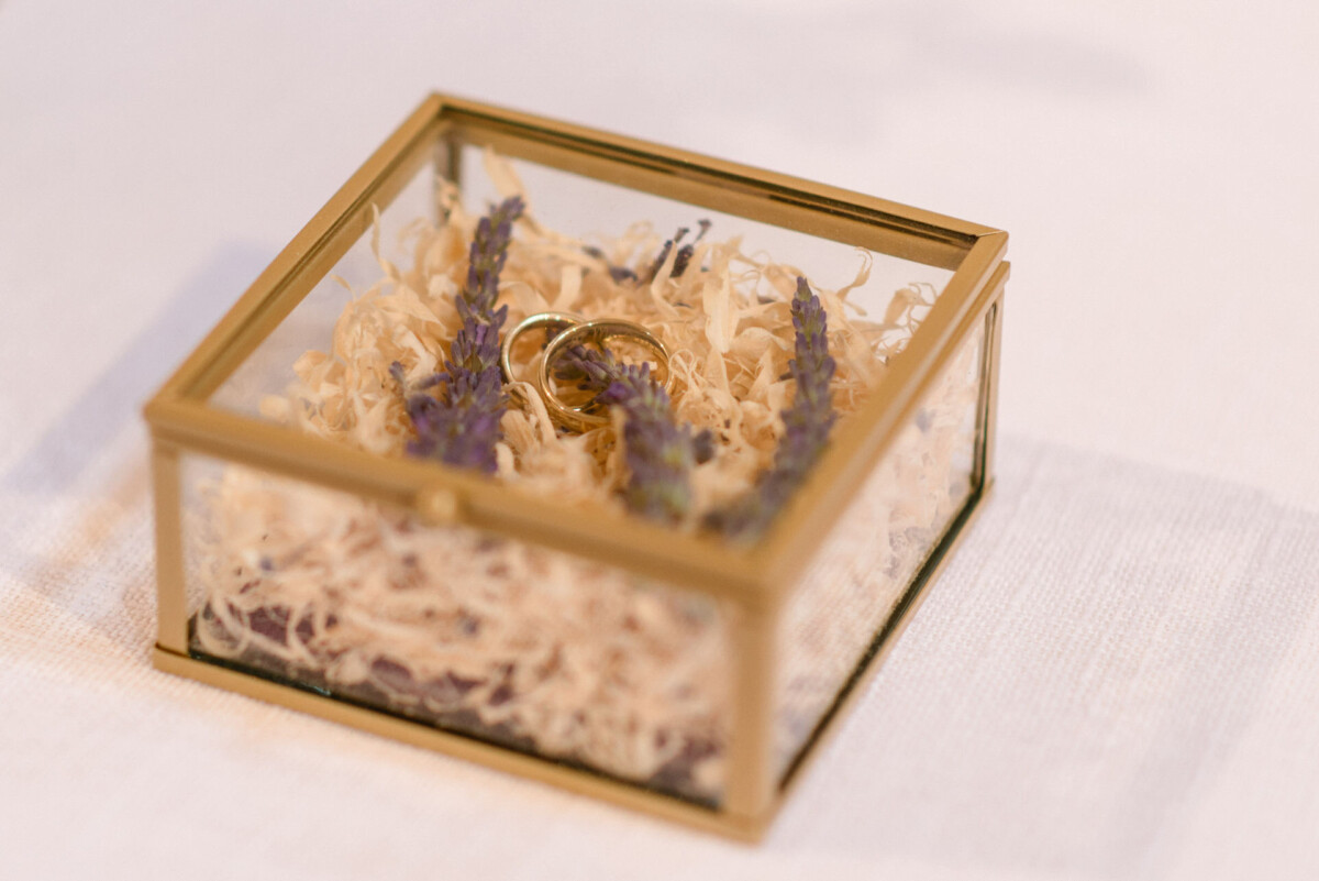 Goldene Ringe in einer Glasbox, geschmückt mit getrockneten lila Blumen und Stroh auf einer weißen Oberfläche.