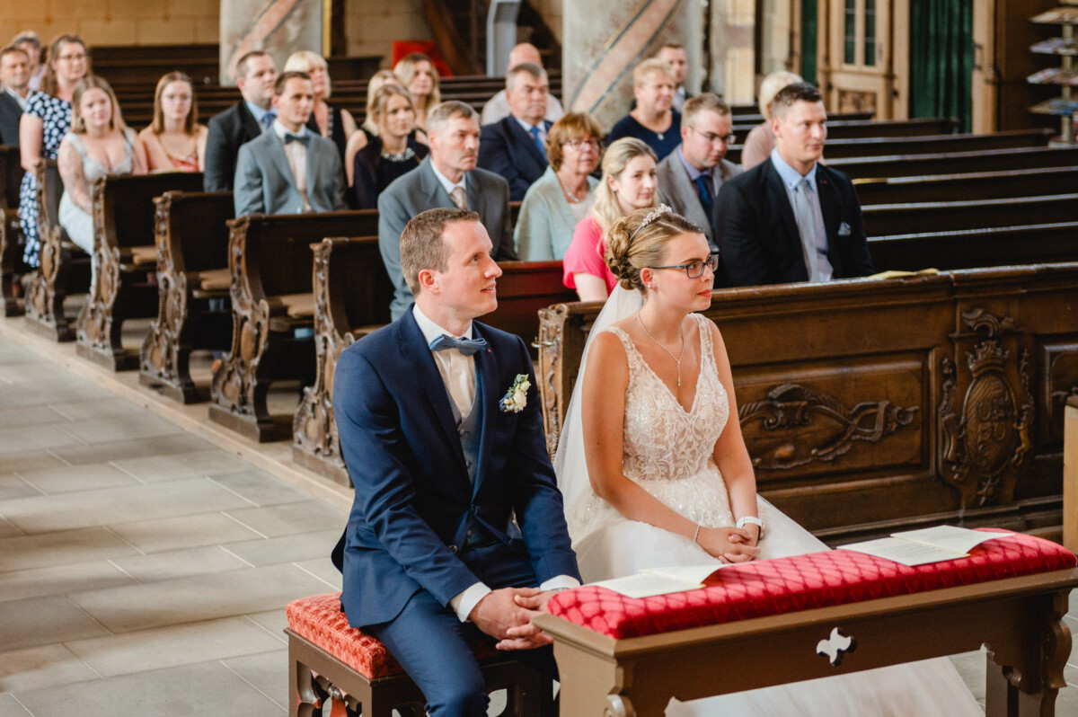 Ein Paar in Abendgarderobe sitzt vor einer Kirche, hinter ihnen sitzen Gäste in den Kirchenbänken.