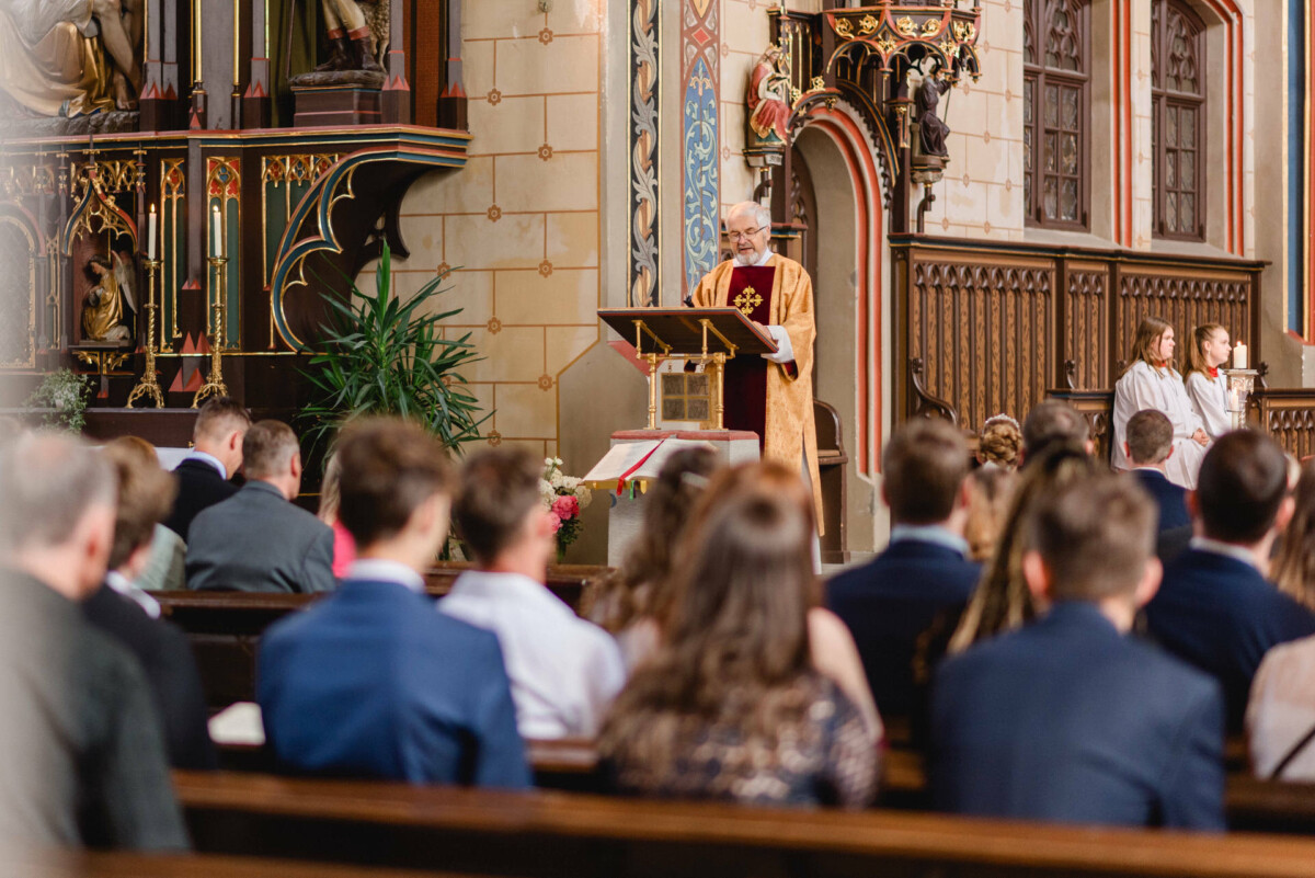 Ein Priester spricht an einem Rednerpult in einer Kirche und wendet sich während eines Gottesdienstes an eine sitzende Gemeinde. Im Inneren der Kirche finden sich kunstvolle Holzschnitzereien und religiöse Ikonen.