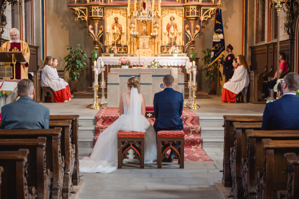 Während einer Hochzeitszeremonie sitzen Braut und Bräutigam in einer Kirche am Altar, umgeben von Priestern und Messdienern.