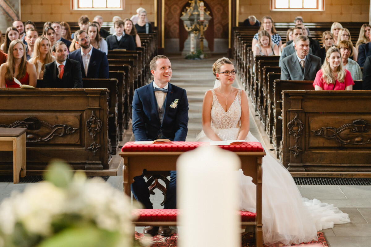 Ein Paar sitzt während einer Hochzeitszeremonie vor einer Kirche, umgeben von Gästen, die in Holzbänken sitzen.