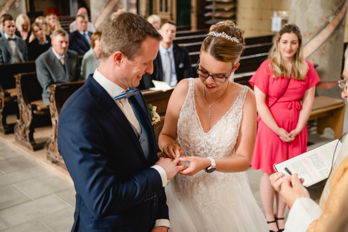 Braut und Bräutigam tauschen bei einer kirchlichen Zeremonie Ringe aus. Im Hintergrund sitzen Gäste. Die Braut trägt ein weißes Kleid, der Bräutigam einen dunklen Anzug.