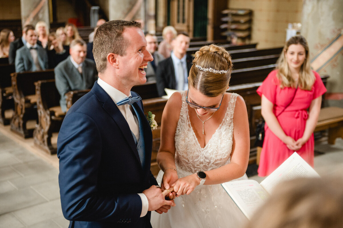 Braut und Bräutigam tauschen während einer Hochzeitszeremonie lächelnd und umgeben von Gästen in einer Kirche Ringe aus.