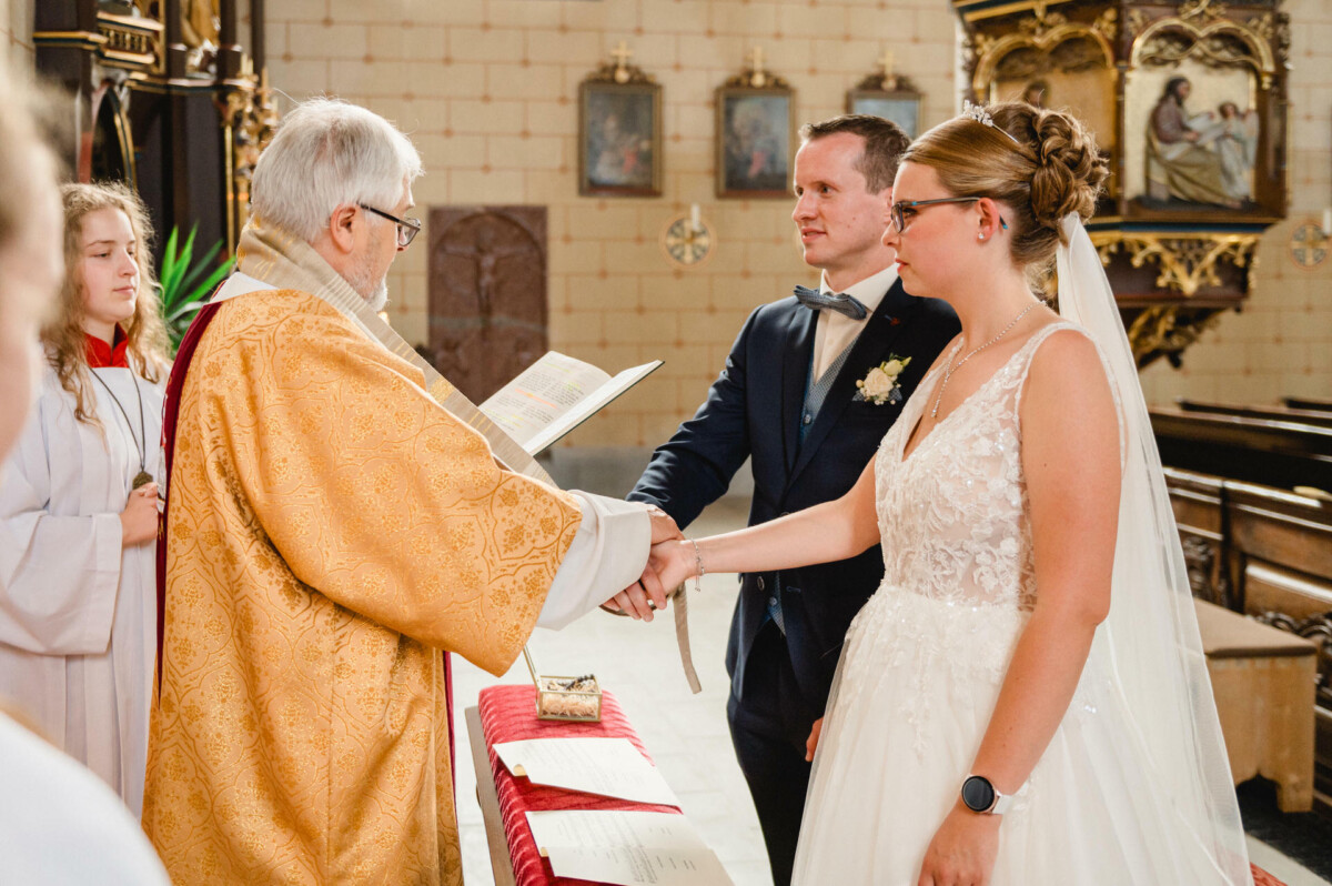 Braut und Bräutigam stehen während einer Hochzeitszeremonie in einer Kirche und halten sich an den Händen. Vor ihnen steht eine Person in zeremonieller Kleidung, die ein Buch hält.