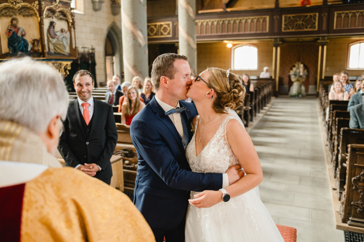 Braut und Bräutigam küssen sich in einer Kirche, umgeben von Gästen, die in den Kirchenbänken sitzen. Eine Person in Robe steht daneben und überwacht die Zeremonie.