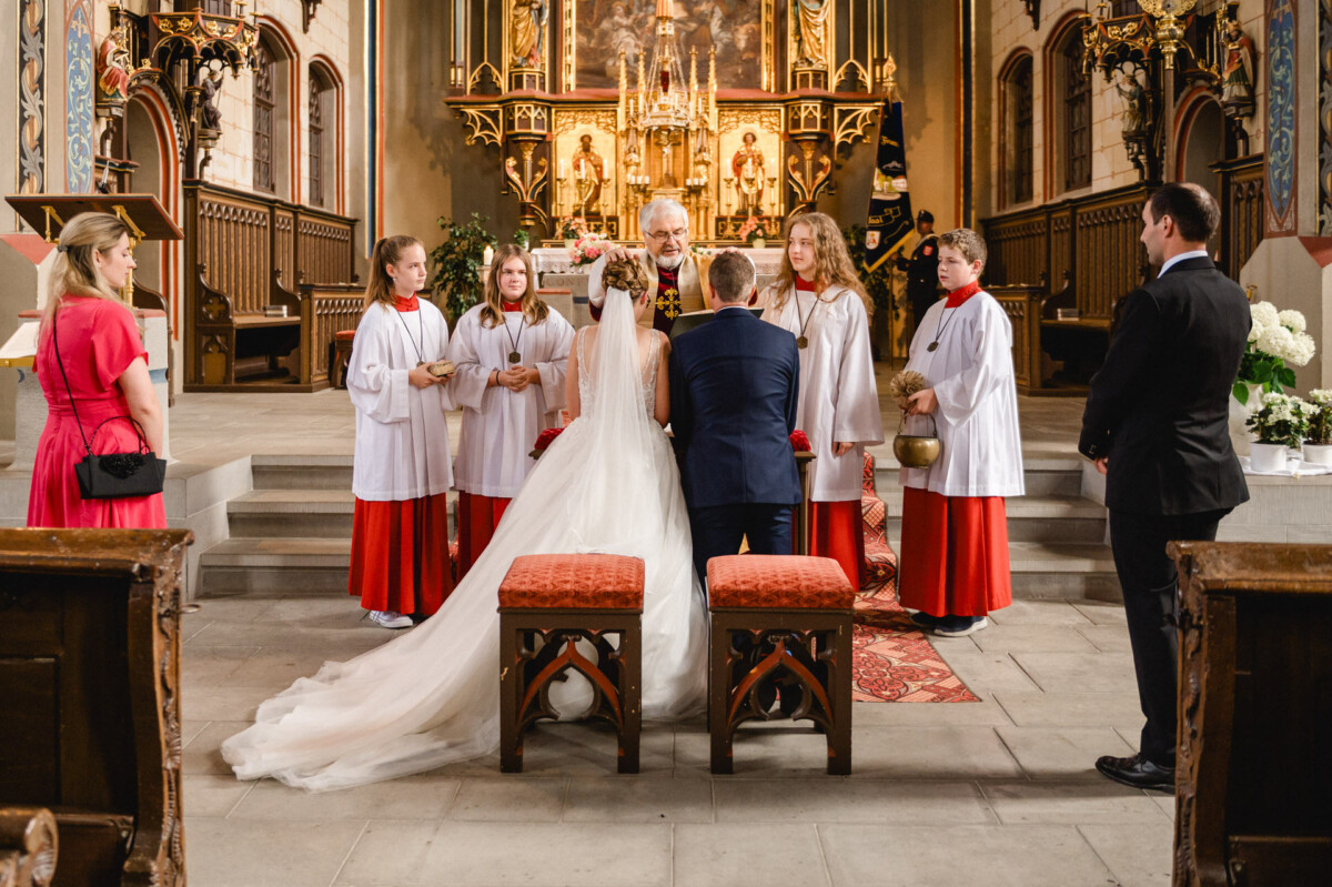 Eine Hochzeitszeremonie in einer Kirche, bei der ein Paar einem Priester gegenübersteht. Mehrere Personen in rot-weißen Gewändern stehen um sie herum.