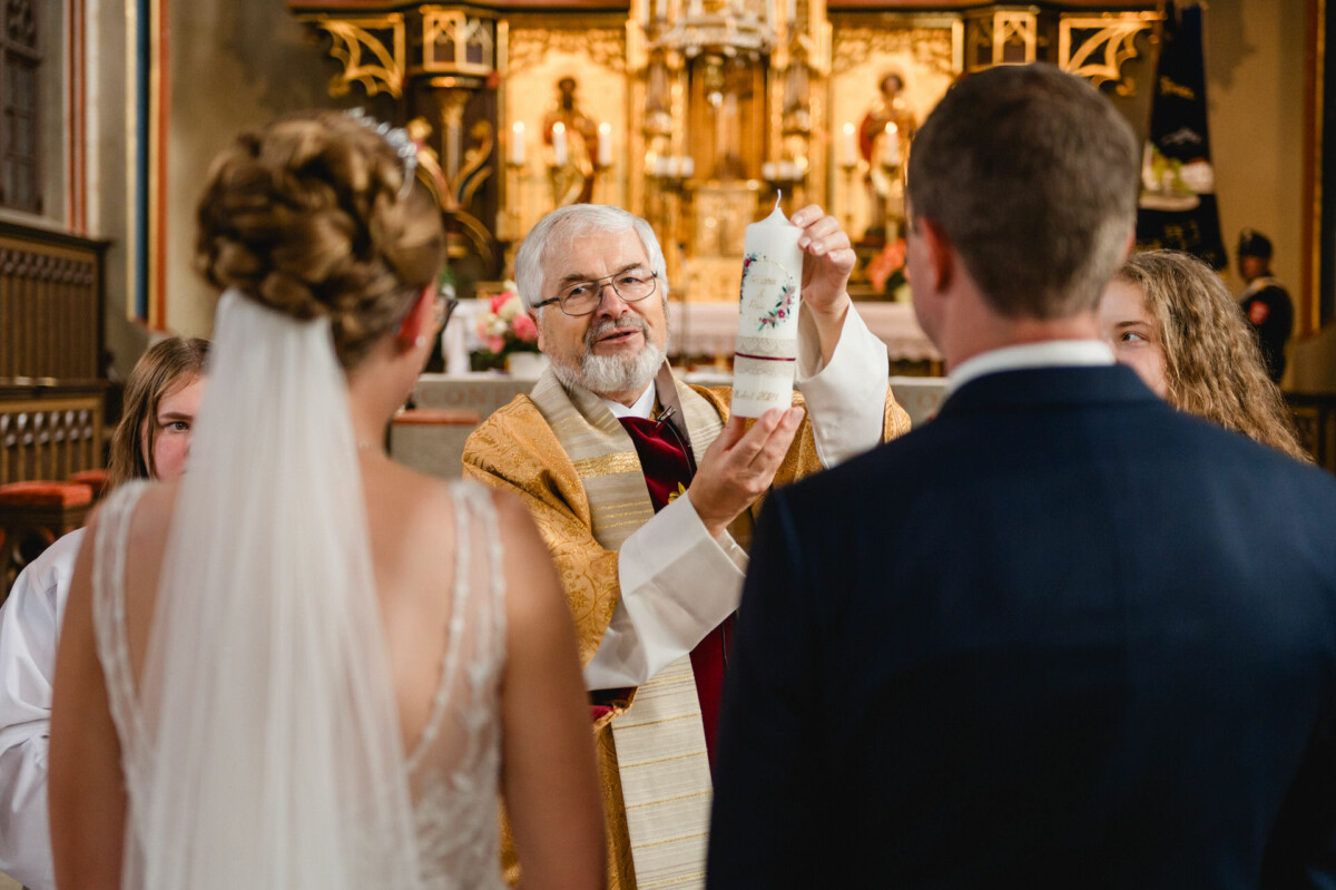 Ein Priester hält während einer Hochzeitszeremonie in einer Kirche eine Kerze hoch, umgeben von Braut, Bräutigam und Gästen.