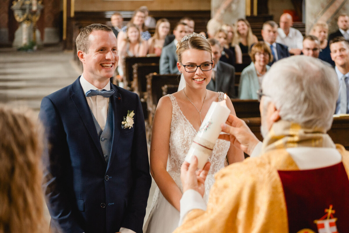 Braut und Bräutigam lächeln, während ein Priester während der Hochzeitszeremonie eine Kerze hält. Im Hintergrund sitzen die Gäste.