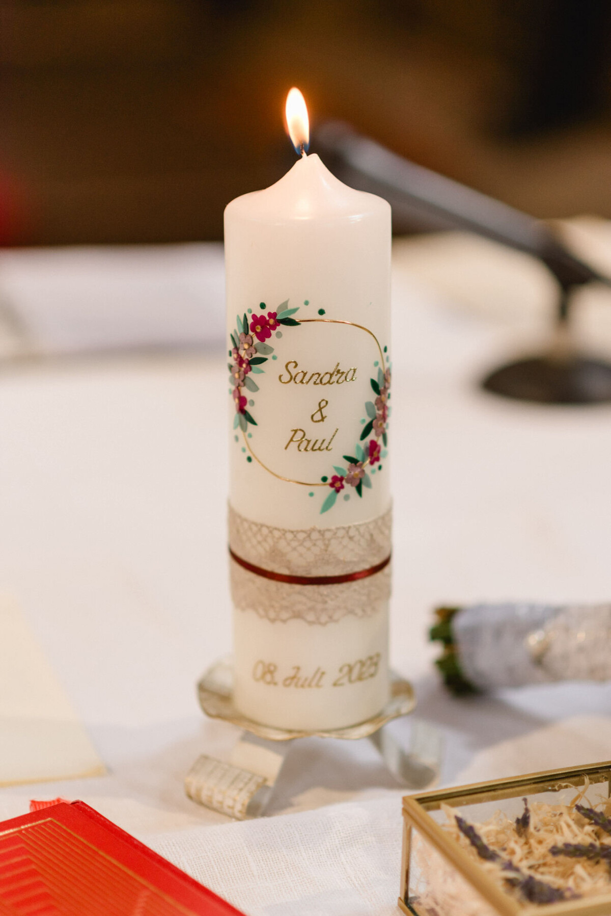 Eine brennende weiße Kerze, verziert mit Blumen und Spitze, mit den Namen „Sandra & Paul“ und dem Datum „08. Juli 2023“, platziert auf einem Halter auf einem Tisch.