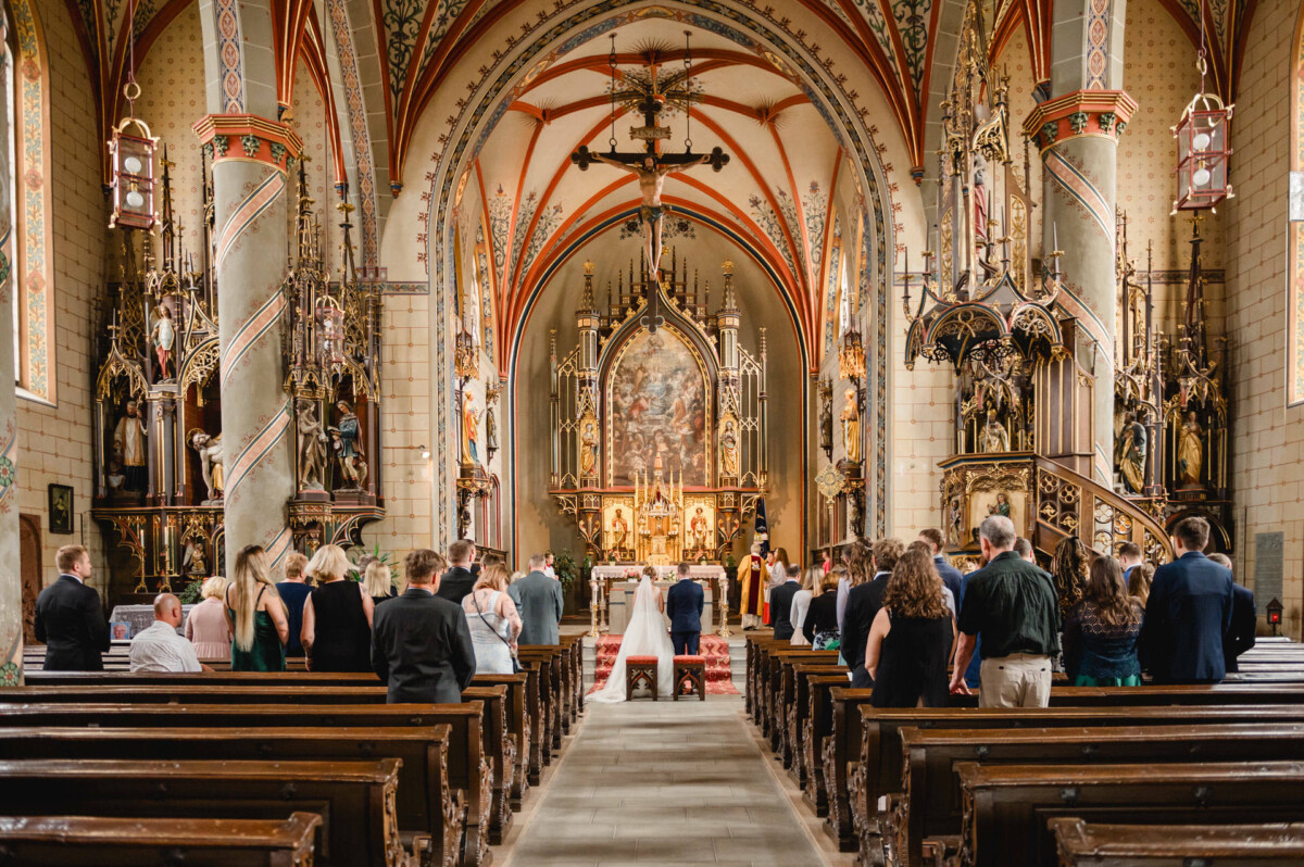 Eine Hochzeitszeremonie in einer Kathedrale mit kunstvoller Architektur und Dekoration. Die Gäste sitzen und ein Paar steht mit dem Geistlichen am Altar.