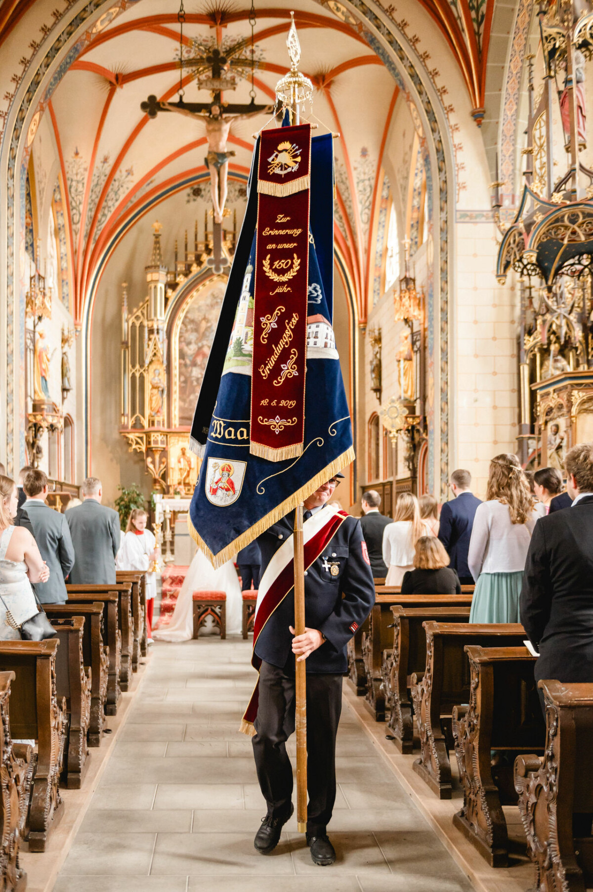 Eine Person in zeremonieller Uniform trägt während einer offiziellen Veranstaltung eine große Flagge den Gang einer reich verzierten Kirche entlang.