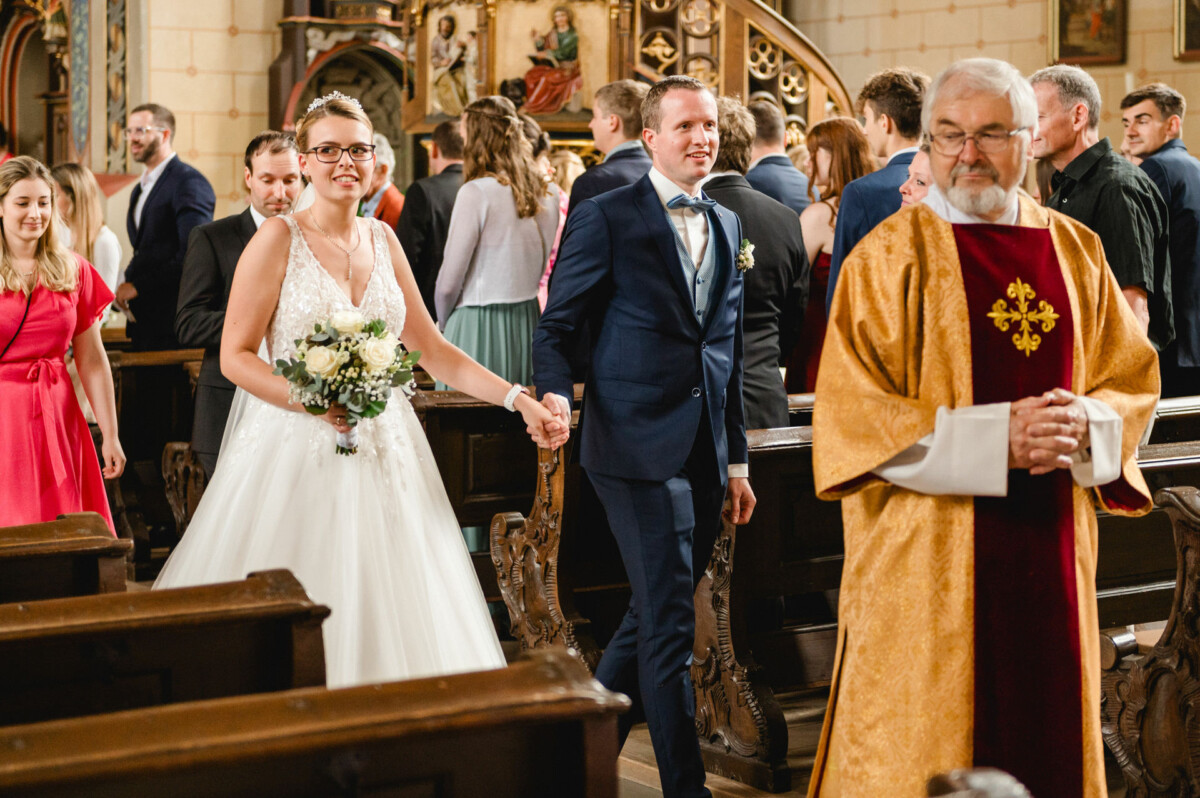Braut und Bräutigam gehen Hand in Hand den Gang in einer Kirche entlang, gefolgt von einem Priester in einer goldenen Robe. Gäste sitzen und stehen.