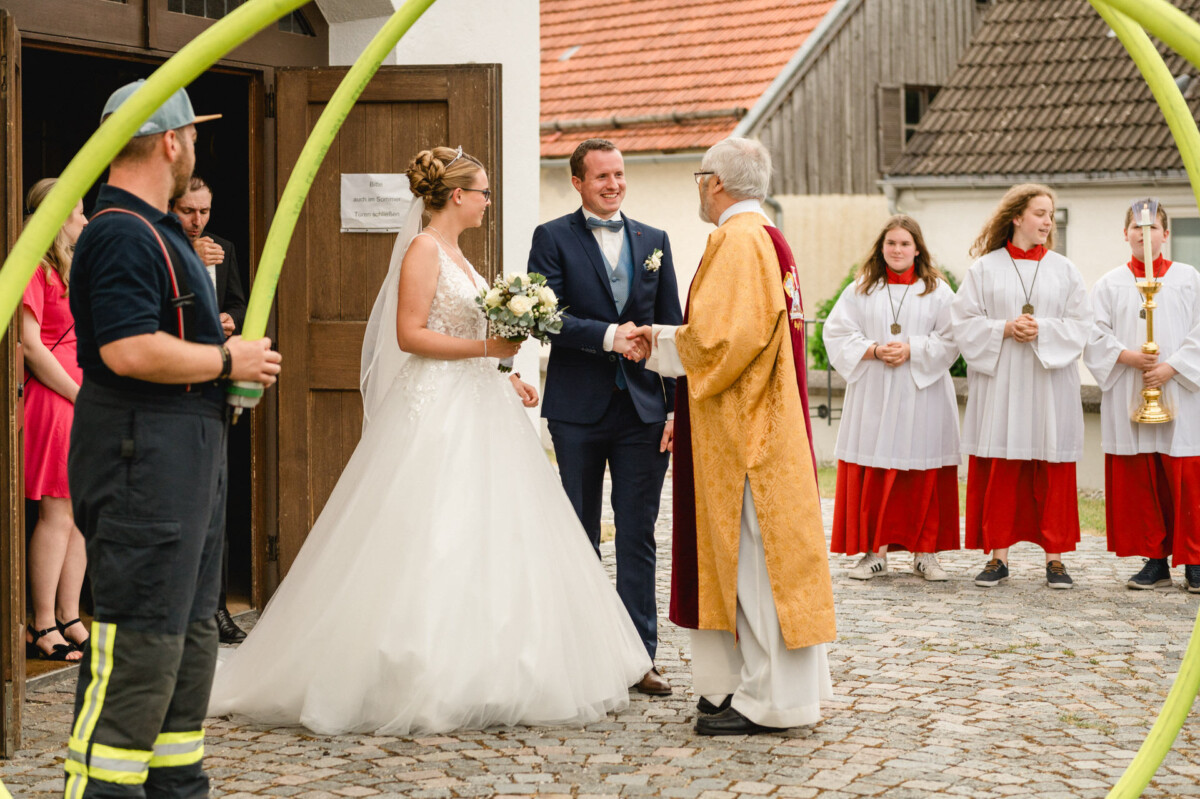 Braut und Bräutigam werden vor einem Gebäude von einem Priester begrüßt. In der Nähe steht eine Gruppe von Menschen in Ordens- und Feuerwehrkleidung.